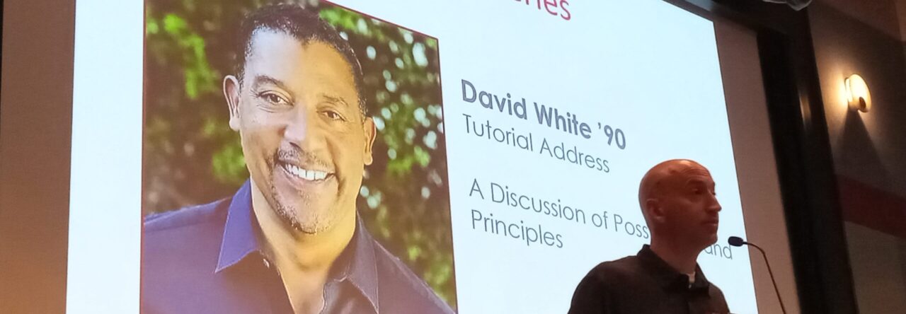 Event host at microphone, with event speaker photo on screen behind the host.