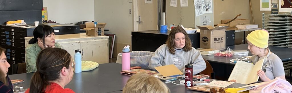 A student shares their comic sketch with the instructor and other students seated around the table. 