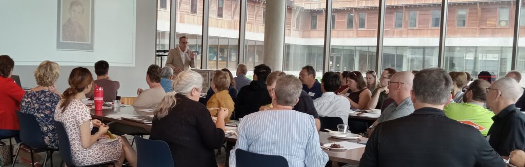 People of different ages sit at tables watching a man in a suit give a talk. 